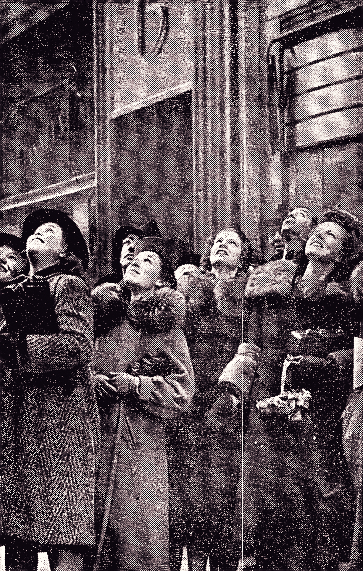 A crowd of people in winter coats look upward expectantly.