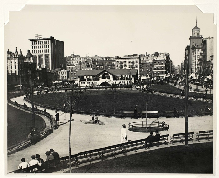 An open lawn is surrounded by a circular pathway and ringed with benches. Brick buildings cluster in the background.