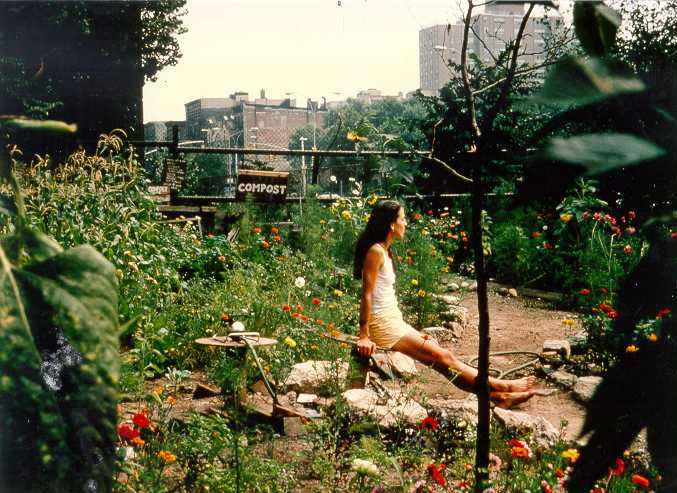 A woman sits in a lush community garden, surrounded by flowers. Around the edges of the frame, multistory buildings hint at the city beyond.