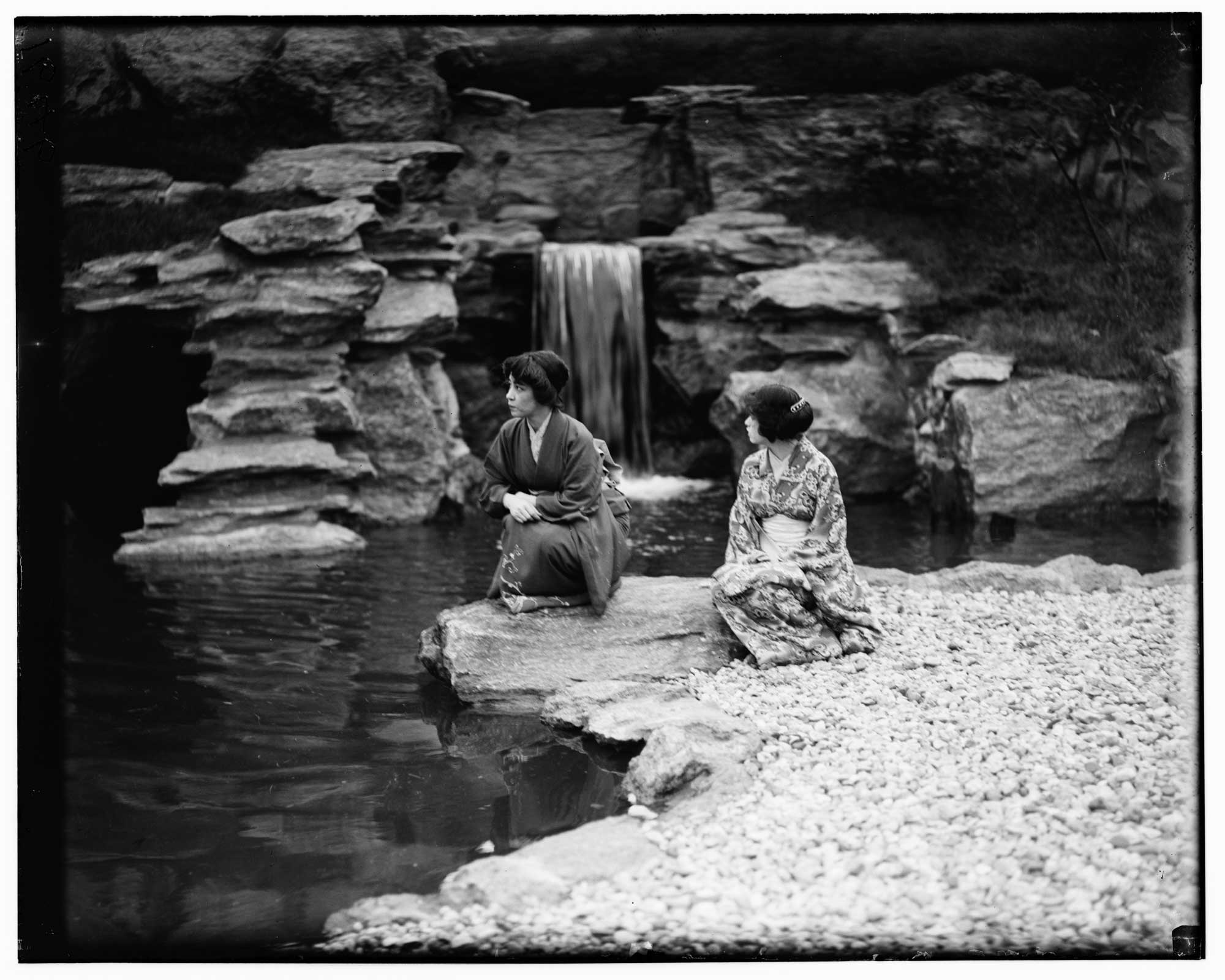 Archival photograph of two Japanese women wearing kimonos in front of a small waterfall.