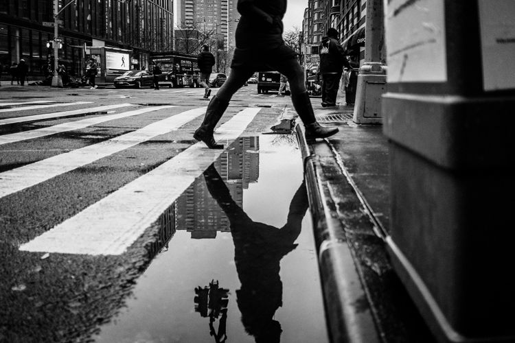 A pedestrian strides across a long gutter puddle on the way to the safety of the sidewalk.