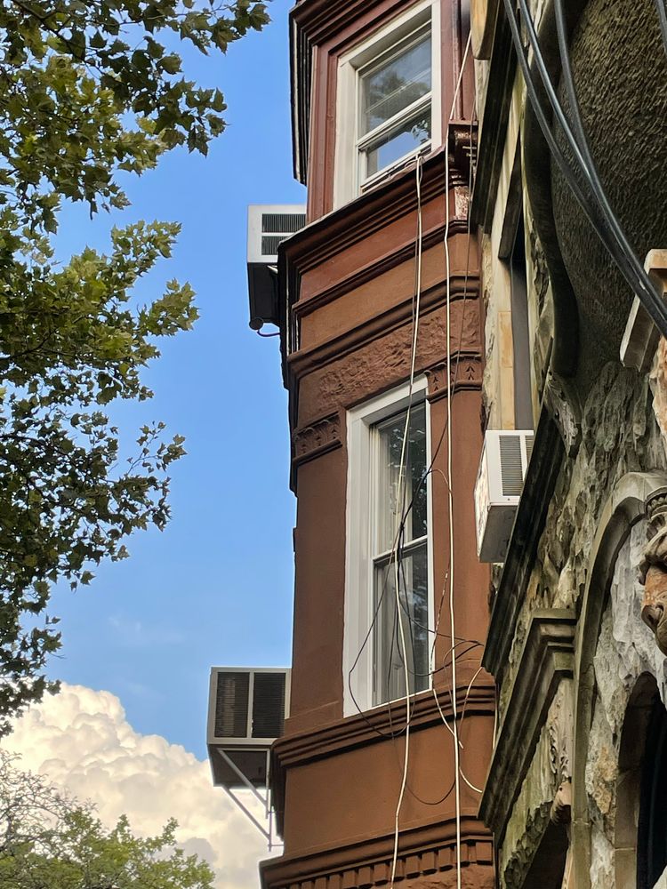 Window air conditioning units jut out in perpendicular cubes from the floors of a brownstone.