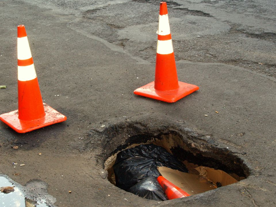 Two traffic cones stand guard before a gaping pothole that has already eaten their comrade cone, as well as some trash.