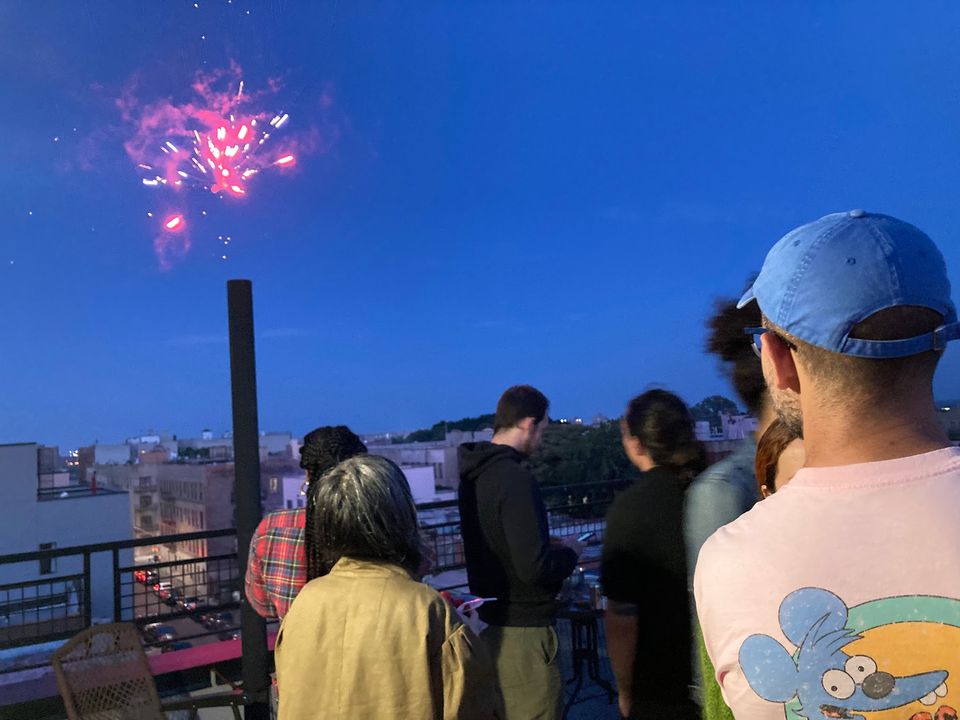 A firework explodes in the twilight sky over a group of people on a Brooklyn rooftop.