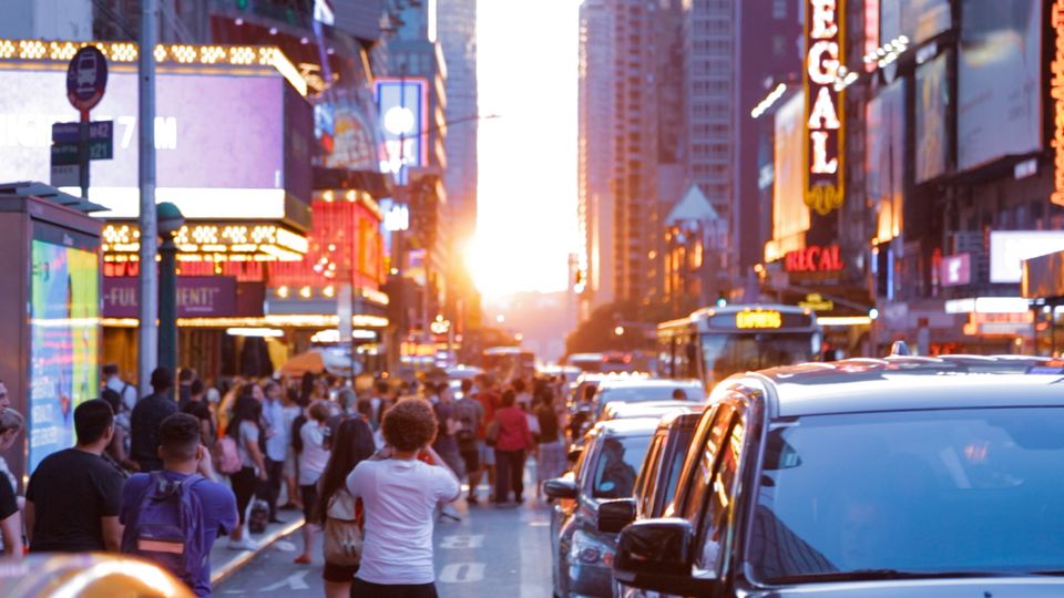 Photo-snapping pedestrians and cars jostle for road space on 42nd street as the Sun flares between buildings.